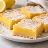 Powdered-sugar lemon bars with golden shortbread crust on a white plate beside a halved lemon.
