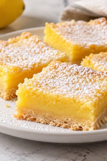 Powdered-sugar lemon bars with golden shortbread crust on a white plate beside a halved lemon.