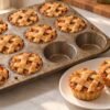 Golden lattice-topped mini apple pies in a muffin pan, with two pies served on a white plate.